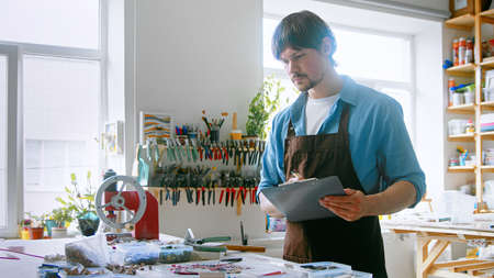 A young master conducting inventory in a creative studio. A man in an apron with clipboard at workの写真素材