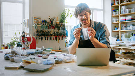 Happy man conducts training online using a laptop in the studio. Young male in an apron talking by video call in a workshopの写真素材