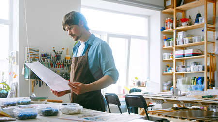Young master in uniform looking at art sketches on the table in the studio. Young adult man at art workの写真素材