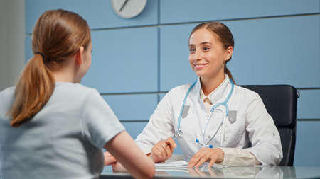 Smiling blonde general practitioner in white coat with blue stethoscope consults young woman with long hair in ponytail sitting at tableの写真素材