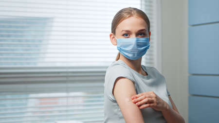 Young woman blonde in blue disposable face mask examines injection spot on forearm and looks straight standing in hospital room close viewの写真素材