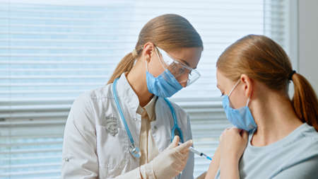 Blonde medical nurse in disposable face mask and glasses fills syringe with anti covid vaccine and injects young woman patient close viewの写真素材