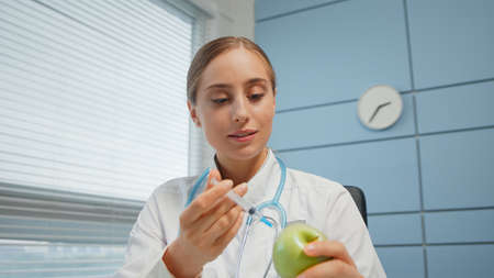 Blonde young woman medical blogger in white coat shows syringe and explains injection process holding fresh green apple close viewの写真素材