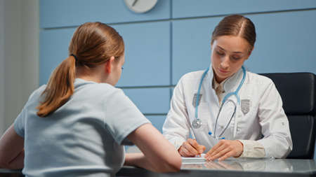 Lady doctor in white coat with stethoscope writes prescription for young woman patient in medical file with pen on hospital tableの写真素材