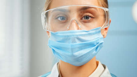 Concentrated lady doctor in blue disposable facial mask and protective goggles looks straight standing in hospital room super close viewの写真素材