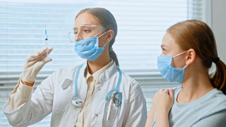 Professional lady general practitioner injects young woman patient in blue disposable face mask against hospital window close viewの写真素材