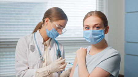 Professional medical nurse in white coat and blue face mask injects young woman patient in arm sitting in hospital room close viewの写真素材