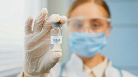 Blurred young woman medical nurse in disposable mask and glasses holds coronavirus vaccine vial in hand with white glove extreme close viewの写真素材