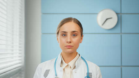 Blonde young woman general practitioner in white coat with blue stethoscope looks straight standing against hospital wall close viewの写真素材