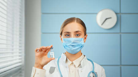Young woman doctor in blue disposable face mask and white coat holds vial with vaccine and looks straight standing in hospital close viewの写真素材