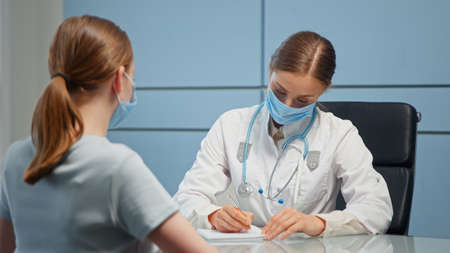 Lady medical consultant in facial mask and white coat examines blonde patient talking and writing in medical file in hospitalの写真素材