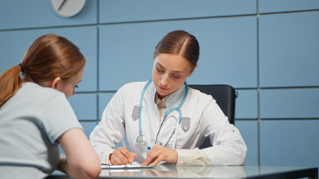 Young woman doctor in white coat examines hospital visitor listening carefully about problems and writing in medical file with penの写真素材