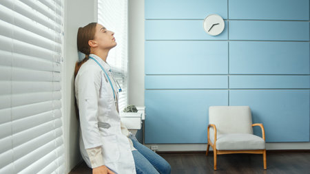 Exhausted young woman doctor in white coat sits down to rest on gray designed windowsill and takes off blue disposable mask in hospital roomの写真素材