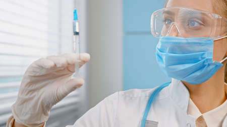 Blonde practitioner in disposable face mask and gloves fills syringe with vaccine against covid holding vial in hospital close viewの写真素材