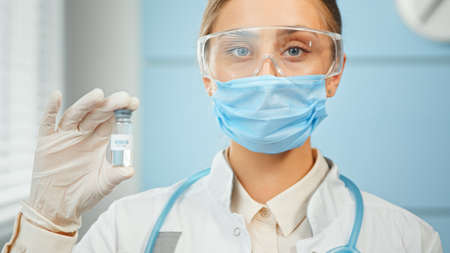 Young woman researcher in blue disposable mask glasses and white coat holds transparent vial with vaccine in laboratory close viewの写真素材