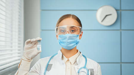 Concentrated young woman doctor in blue disposable face mask and glasses holds vaccine vial in hand with white sterile glove closeupの写真素材