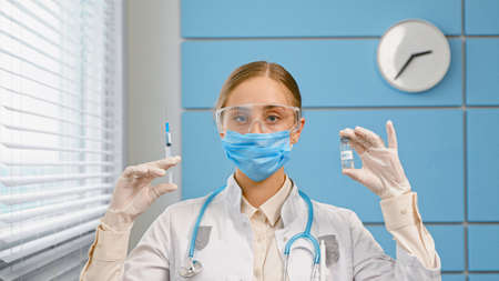 Young woman blonde in white coat and mask with stethoscope holds syringe and vial in hands with sterile gloves in hospital close viewの写真素材