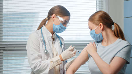 Blonde lady hospital nurse in disposable facial mask and glasses injects young woman patient arm sitting in clinic office close viewの写真素材