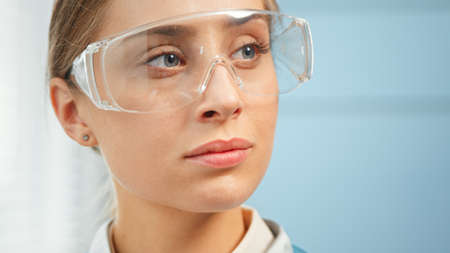 Exhausted young woman medical nurse in protective glasses looks straight standing in hospital room under bright sunlight close viewの写真素材