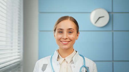 Young woman general practitioner in white coat with blue stethoscope looks straight standing against hospital wall close viewの写真素材