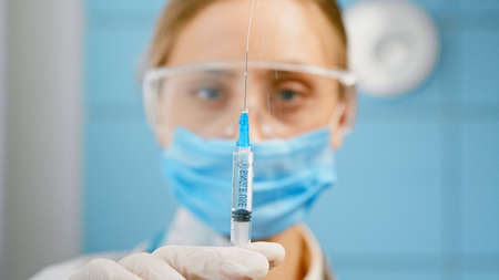 Blurred blonde nurse in blue disposable face mask and glasses presses syringe and sprays vaccine standing in hospital close viewの写真素材
