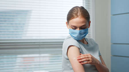Young girl in blue disposable face mask examines injection spot on forearm in hospital room close viewの写真素材