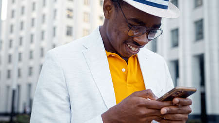 Joyful black man in glasses and suit types on smartphone standing on city street among local highrise office buildings close viewの写真素材