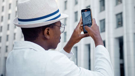 Concentrated black guy in classic suit films highrise office buildings holding smartphone under bright sunlight close viewの写真素材