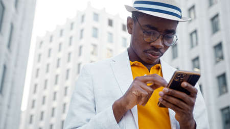 Black man in suit with hat types on smartphone and laughs cheerfully standing by office buildings under bright sunlight close viewの写真素材