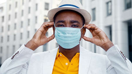 Serious African American guy in suit and hat looks straight and puts blue disposable face mask on against city building closeupの写真素材