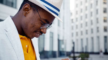 Concentrated African American man in glasses and hat looks at smartphone display and types standing against buildings closeupの写真素材