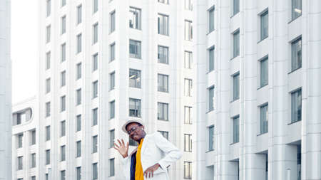 Amazed black guy in yellow t shirt and white suit dances contemporary dance against megalopolis office buildings lit by sunlightの写真素材