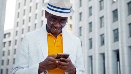 Smiling African American man in glasses and hat types on smartphone and smiles with happiness against blurred highrise buildings closeupの写真素材