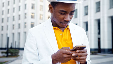 Concentrated black guy in t shirt and stylish suit with fedora hat types on smartphone and smiles cheerfully on city square closeupの写真素材