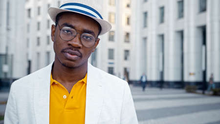 Serious black man in classic suit and glasses looks straight standing on gray paved street with highrise office buildings closeupの写真素材