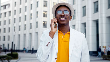 African American businessman in white suit and fedora hat talks on smartphone walking along megalopolis street lit by sunlight closeupの写真素材