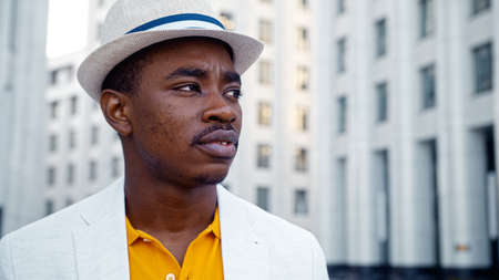 Serious African American man in white suit with fedora hat looks around standing on empty street against buildings close viewの写真素材