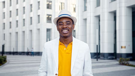 Smiling African American businessman in white fedora hat with blue strip walks along city square with highrise city buildings closeupの写真素材