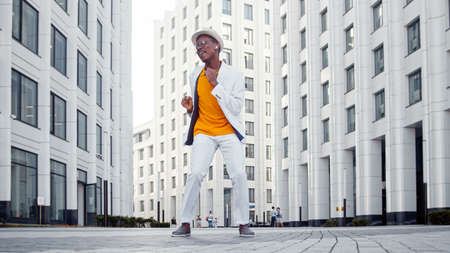 Black guy in orange t shirt under white jacket with hat and sunglasses dances on gray paved road among large megalopolis buildingsの写真素材