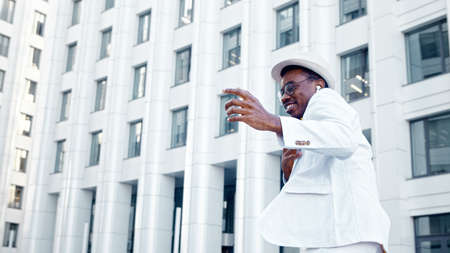 Amazed black man in white suit with hat and glasses listens to music in wireless headphones and dances on street against building closeupの写真素材