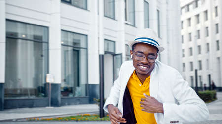 Amazed black guy in yellow t shirt and white suit dances and sings on empty gray street pavement among city buildings under sunlightの写真素材