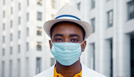 American man in blue disposable face mask looks straight standing on street against highrise city building close viewの写真素材