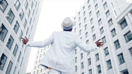 Afro American guy in white suit and Fedora hat walks along gray pavement dancing and listening to music in headphones backside viewの写真素材