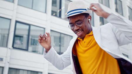 African American man in hat and wireless headphones listens to music and dances with joyful smile on megalopolis street close viewの写真素材