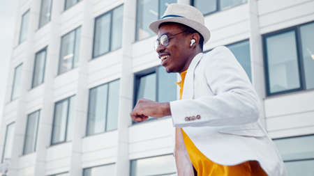 Smiling guy listens to music in wireless headphones and dances on street with buildings under bright sunlight low angle shotの写真素材