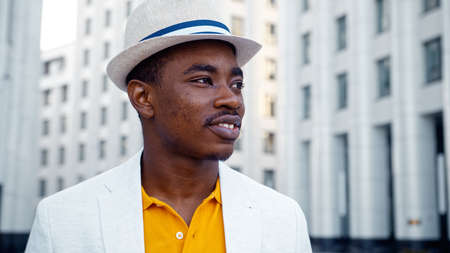 Thoughtful man in yellow t shirt and white classic suit with hat looks around standing on street with highrise buildings closeupの写真素材