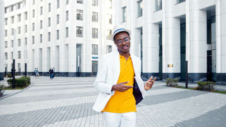 Joyful black man in glasses and white wireless headphones listens to music and dances walking along gray pavement in megalopolisの写真素材