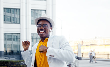 Inspired black actor in stylish suit dances and sings on city square with highrise buildings under sunlight close low angle shotの写真素材
