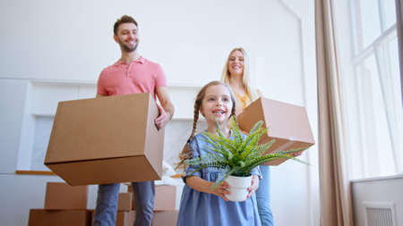 Delighted family parents and preschooler daughter move to new apartment holding brown cardboard boxes and pot plant low angle shotの写真素材