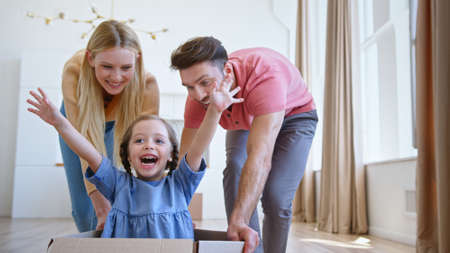 Joyful parents play with little funny daughter in blue dress moving kid along new apartment floor in large brown cardboard boxの写真素材
